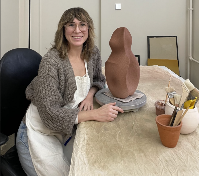 Artist Yasha Butler sits at her work table in her studio, smiling warmly at the camera. She wears glasses, a gray knit cardigan over a white apron, and has shoulder-length hair with bangs. On the table is a terracotta-colored ceramic sculpture on a gray wheel, alongside pottery tools in a clay pot and other studio supplies. The workspace appears bright and organized, with framed artwork visible in the background.
